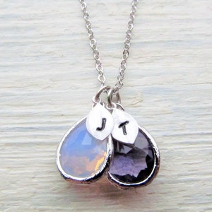 May include: A silver chain necklace with two teardrop-shaped pendants. The pendants are made of clear glass with a light purple and a light pink hue. The pendants have silver charms with the letters "J" and "T" on them.