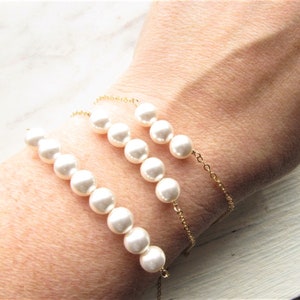 May include: Three gold chain bracelets with white pearl beads. The bracelets are layered on a person's wrist.