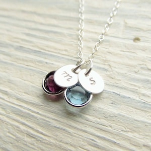 May include: A silver necklace with two small charms, one with the letter "M" and the other with the letter "S". The charms are attached to a silver chain and have small gemstones, one purple and one blue.