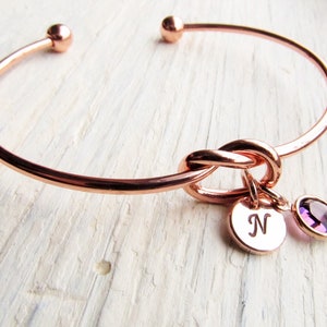 May include: Rose gold bangle bracelet with a knot design and a charm with the letter N and a purple gemstone.