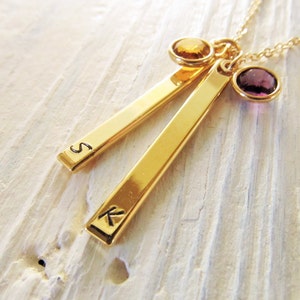 May include: Gold necklace with two bar pendants engraved with the letters "S" and "K". The necklace features a purple and a yellow gemstone.