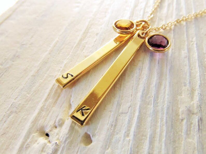 May include: Gold necklace with two bar pendants engraved with the letters "S" and "K". The necklace features a purple and a yellow gemstone.