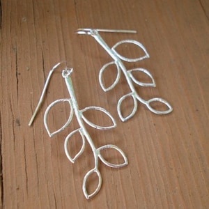 May include: A pair of silver earrings shaped like a branch with leaves. The earrings are delicate and have a simple design.