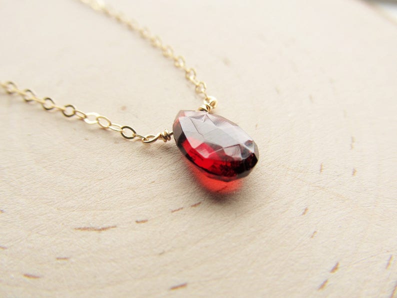 May include: A delicate gold chain necklace with a single, faceted, red gemstone pendant.