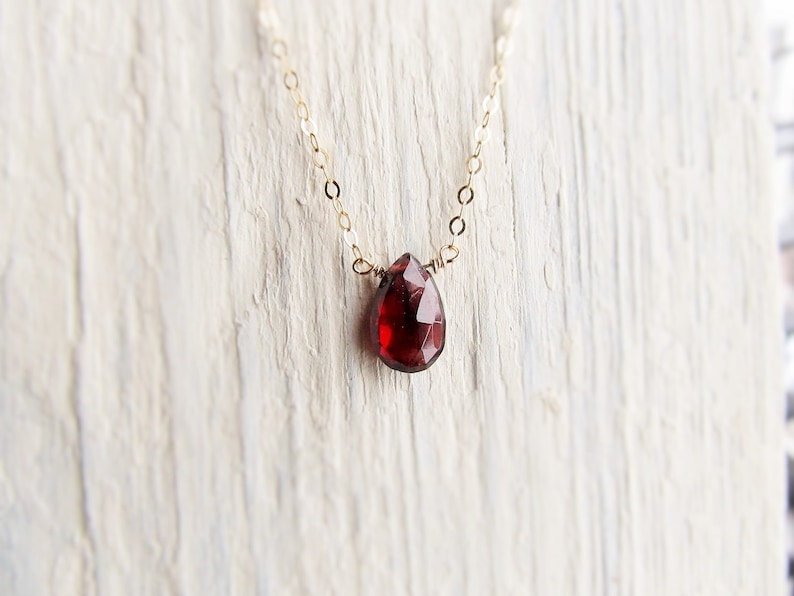 May include: A delicate gold chain necklace with a single teardrop-shaped red gemstone pendant.