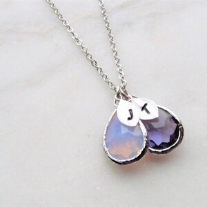 May include: A silver chain necklace with two teardrop-shaped pendants. One pendant is clear with a light pink hue, and the other is a dark purple. Both pendants have silver leaf charms with the initials "J" and "K" on them.