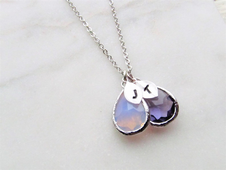 May include: A silver chain necklace with two teardrop-shaped pendants. One pendant is clear with a light pink hue, and the other is a dark purple. Both pendants have silver leaf charms with the initials "J" and "K" on them.