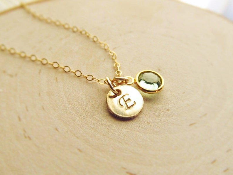 May include: A gold chain necklace with a small gold disc charm with the letter "E" engraved on it. A small green gemstone is attached to the charm.
