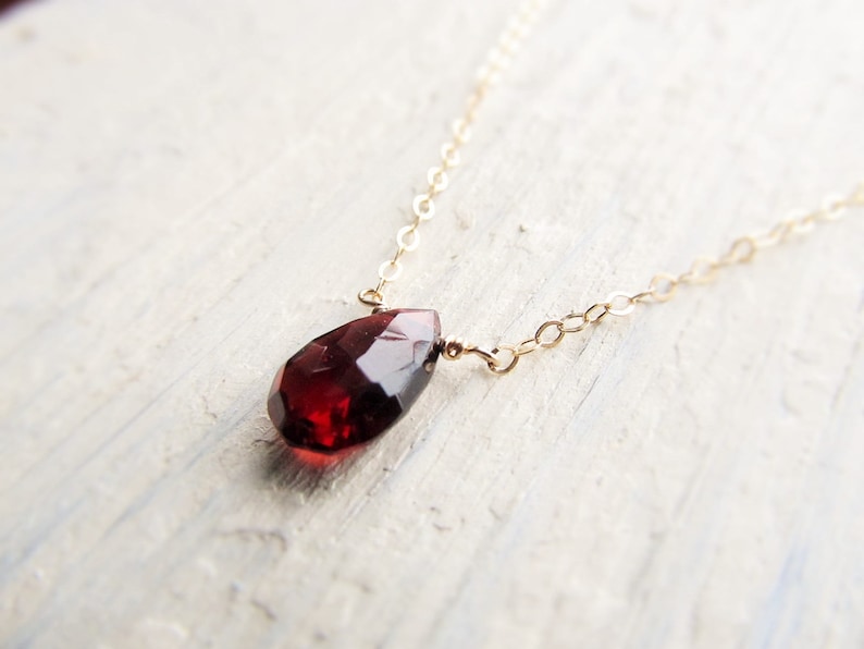 May include: A delicate gold chain necklace with a single, teardrop-shaped red garnet pendant.