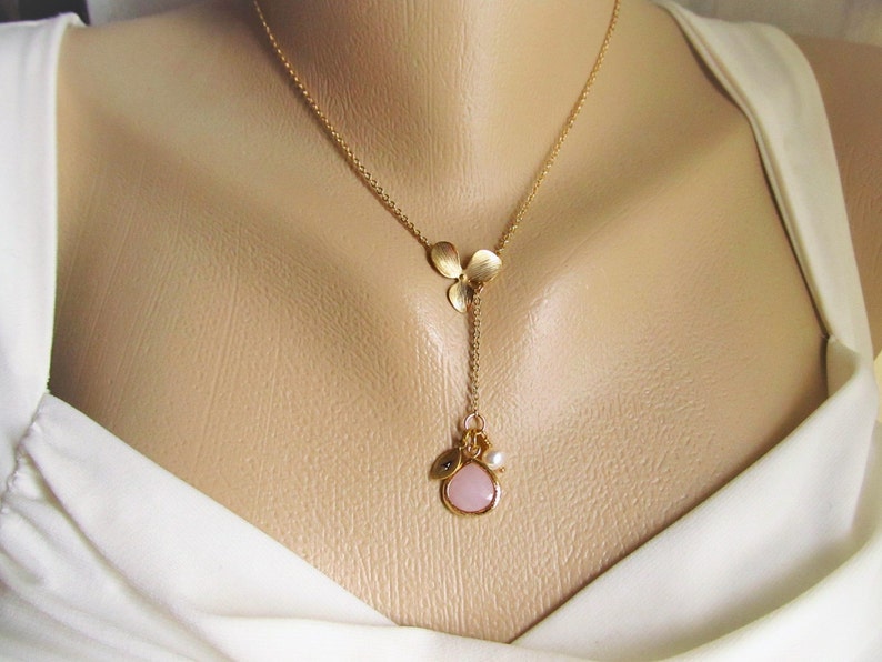 May include: A gold chain necklace with a pink stone pendant and a charm with three gold leaves. The necklace is on a white mannequin.