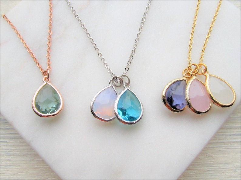 May include: Three delicate necklaces with teardrop-shaped pendants. The necklaces are made of gold, silver, and rose gold chains. The pendants are made of various colored gemstones, including green, blue, purple, pink, and white.