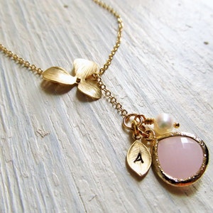 May include: A gold necklace with a delicate flower charm, a small pink gemstone pendant, and a pearl accent. The pendant has a small gold leaf charm with the letter 'A' on it.