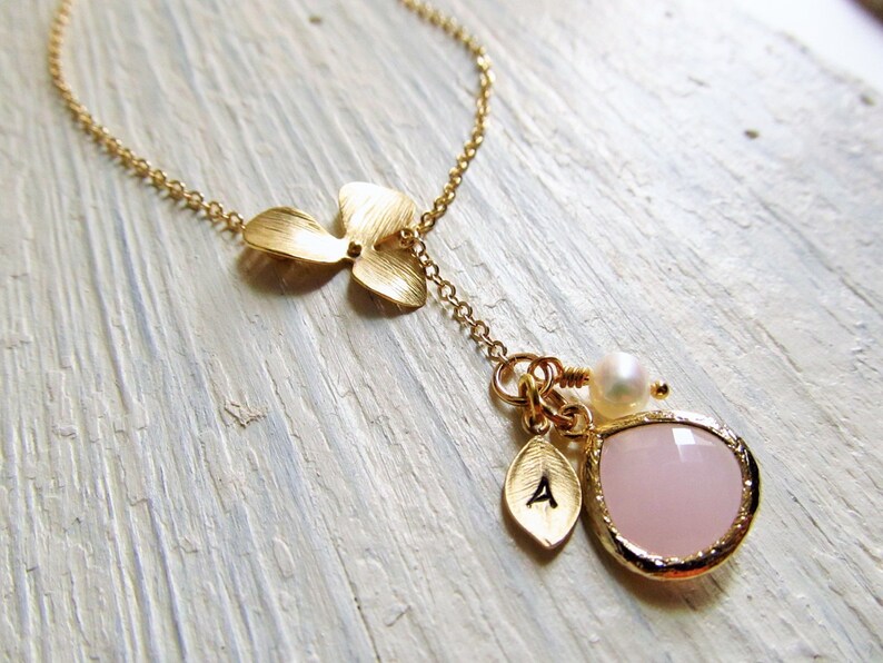 May include: A gold necklace with a delicate flower charm, a small pink gemstone pendant, and a pearl accent. The pendant has a small gold leaf charm with the letter 'A' on it.