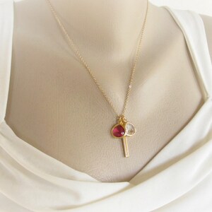 May include: A gold necklace with a pendant. The pendant has a red gemstone and a clear gemstone, plus a small gold bar. The necklace is displayed against a white background.