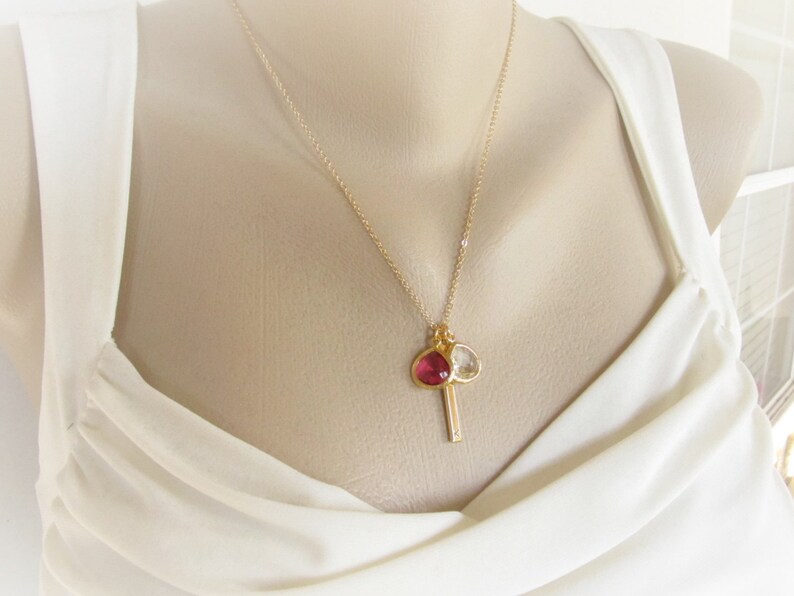May include: A gold necklace with a pendant. The pendant has a red gemstone and a clear gemstone, plus a small gold bar. The necklace is displayed against a white background.