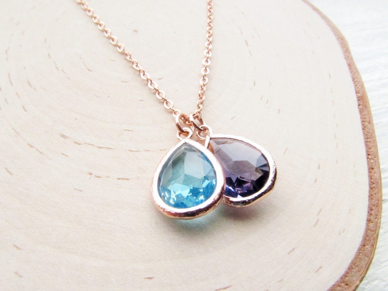 May include: A delicate gold necklace with two teardrop-shaped pendants. The larger pendant is a light blue color, while the smaller pendant is a deep purple.