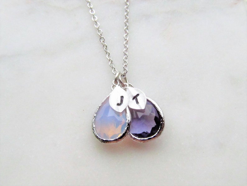 May include: A silver chain necklace with two teardrop-shaped pendants. The pendants are made of clear glass with a light purple hue. The pendants are engraved with the letters "J" and "T".