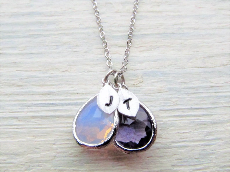 May include: A silver chain necklace with two teardrop-shaped pendants. The pendants are made of clear glass with a light purple and a light pink hue. The pendants have silver charms with the letters "J" and "T" on them.