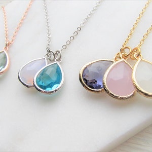 May include: Three silver necklaces with teardrop-shaped pendants. The pendants are made of clear, blue, and purple glass. The necklaces are on a white surface.