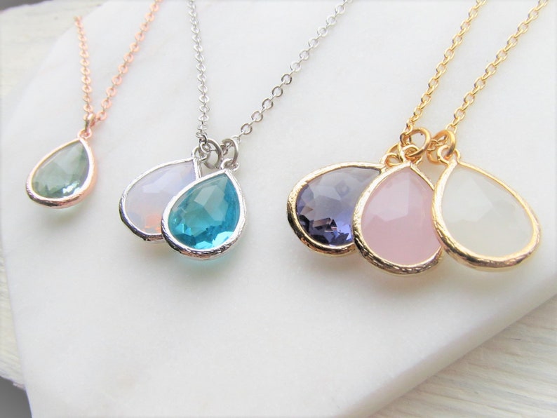 May include: Three silver necklaces with teardrop-shaped pendants. The pendants are made of clear, blue, and purple glass. The necklaces are on a white surface.