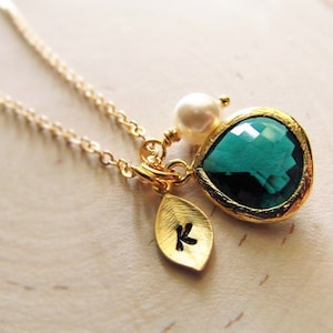 May include: A gold necklace with a teardrop-shaped green gemstone pendant, a small pearl charm, and a leaf charm with the letter "K" engraved on it.