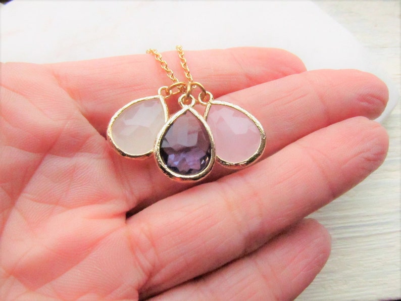May include: A gold chain necklace with three teardrop-shaped pendants. The pendants are made of clear, pink, and purple glass.