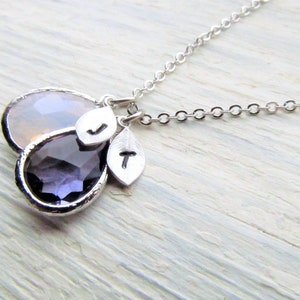 May include: A silver chain necklace with two teardrop-shaped pendants. The larger pendant is a clear, iridescent stone, and the smaller pendant is a deep purple stone. The necklace also features two small silver charms with the letters "J" and "T" on them.