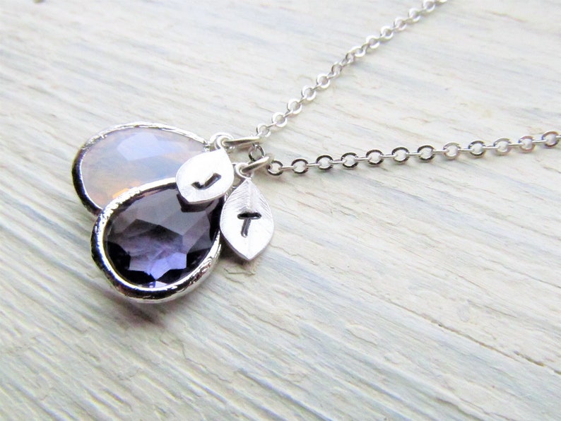 May include: A silver chain necklace with two teardrop-shaped pendants. The larger pendant is a clear, iridescent stone, and the smaller pendant is a deep purple stone. The necklace also features two small silver charms with the letters "J" and "T" on them.
