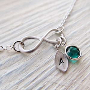 May include: Silver infinity necklace with a small leaf charm and a teal gemstone. The leaf charm has the letter 'A' stamped on it.