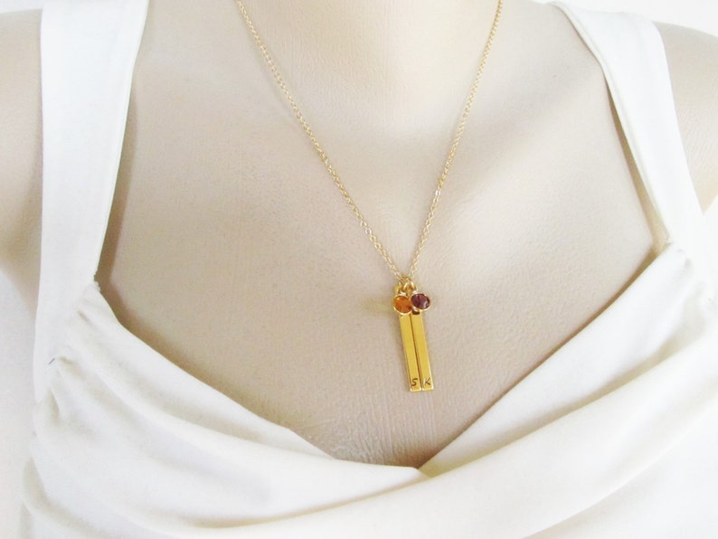 May include: A gold necklace with two small gold bars hanging from it. The bars have the letters "S" and "K" engraved on them. The necklace has two small gemstones, one orange and one purple, attached to the bars.