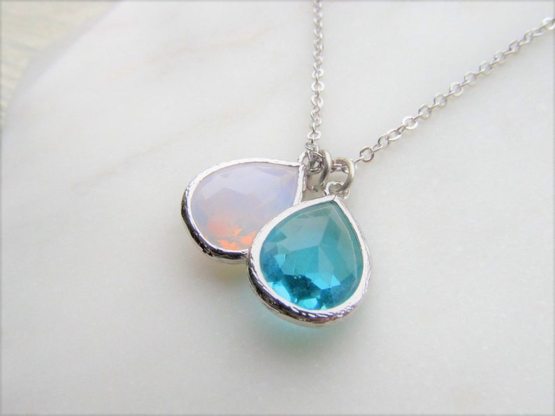 May include: A silver chain necklace with two teardrop-shaped pendants. The top pendant is a clear, iridescent opal, and the bottom pendant is a bright turquoise blue.