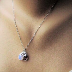 May include: A silver chain necklace with two teardrop-shaped pendants. One pendant is a clear stone, and the other is a purple stone. Both pendants have a small letter stamped on them.