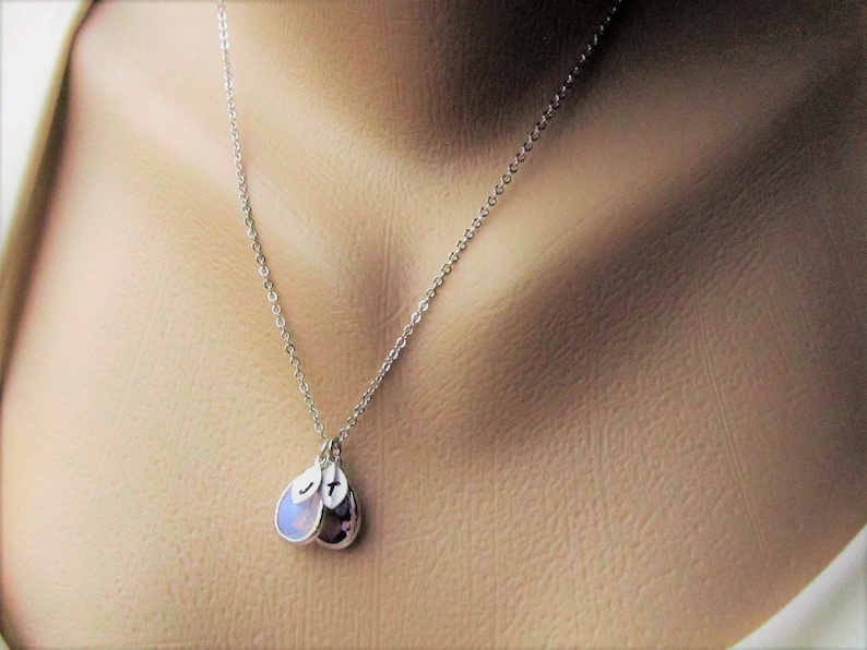 May include: A silver chain necklace with two teardrop-shaped pendants. One pendant is a clear stone, and the other is a purple stone. Both pendants have a small letter stamped on them.