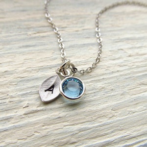 May include: A silver necklace with a blue gemstone and a silver leaf charm with the letter A engraved on it.
