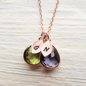 May include: A rose gold necklace with two teardrop-shaped pendants. The pendants are made of clear glass with a green and purple hue. Each pendant has a small rose gold leaf charm with the letters "S" and "N" engraved on them.
