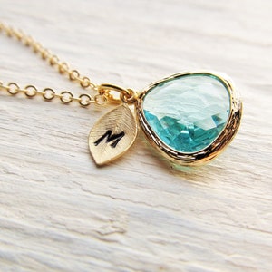 May include: Gold necklace with a teardrop-shaped light blue crystal pendant and a small gold leaf charm with the letter "M" engraved on it.
