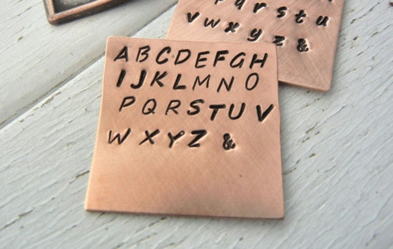 May include: Two copper squares with stamped letters. One features the uppercase alphabet, the other the lowercase. The letters are in a bold, sans-serif font. The copper has a warm, reddish-brown hue. Ideal for personalised crafts or jewellery.