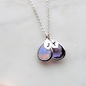May include: A silver chain necklace with two teardrop-shaped pendants. The pendants are made of clear and purple glass and have the letters "J" and "K" engraved on them.