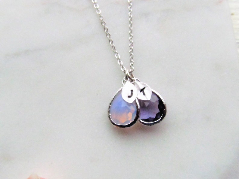 May include: A silver chain necklace with two teardrop-shaped pendants. The pendants are made of clear and purple glass and have the letters "J" and "K" engraved on them.