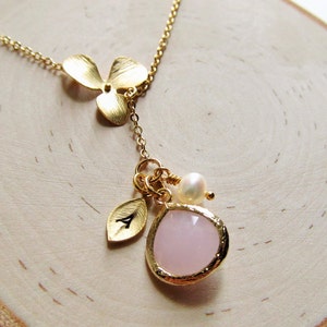 May include: A gold necklace with a delicate flower charm, a pink teardrop-shaped stone, a small leaf charm with the letter A, and a pearl bead.