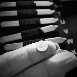 May include: A silver charm with the letter "M" engraved on it. The charm is being held by a hand, and in the background, there are several other charms with letters on them.