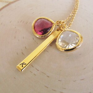 May include: A gold necklace with a bar pendant engraved with the letter "K" and two gemstone charms. One is a teardrop-shaped, deep red stone, the other a clear, faceted stone. The necklace is presented on a light-coloured wooden surface.