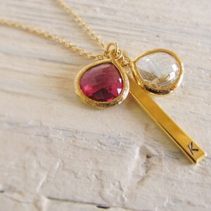 May include: A gold necklace with a fine chain. The pendant includes a teardrop-shaped, deep red gemstone and a clear, teardrop-shaped gemstone, both set in gold. A gold bar with the letter "K" is also part of the pendant.