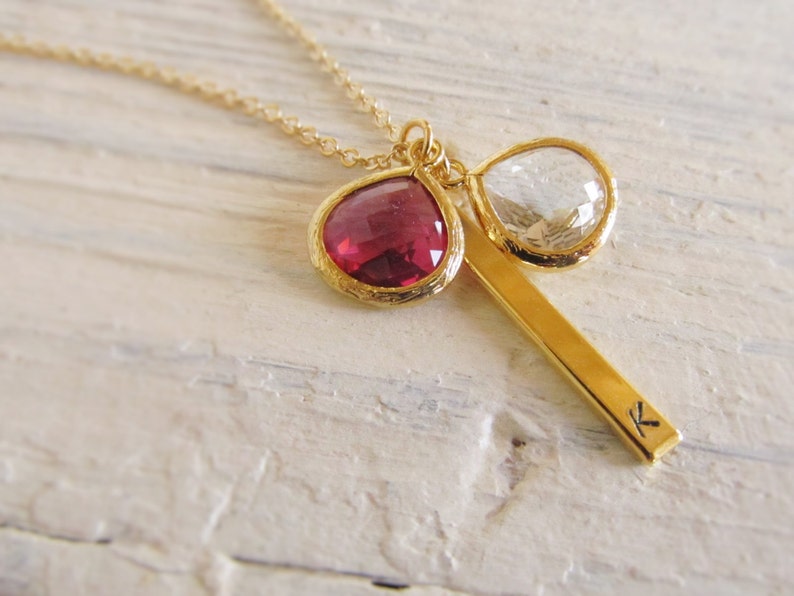 May include: A gold necklace with a fine chain. The pendant includes a teardrop-shaped, deep red gemstone and a clear, teardrop-shaped gemstone, both set in gold. A gold bar with the letter "K" is also part of the pendant.