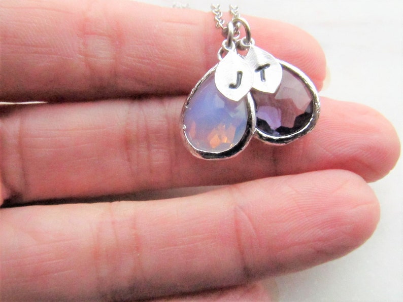 May include: A silver necklace with two teardrop-shaped pendants. The pendants are made of clear and purple glass and have silver initials "J" and "T" on them.