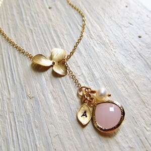 May include: Gold necklace with a delicate flower charm and a pink gemstone pendant. The pendant is accented with a small pearl and a gold letter charm.