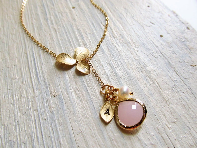 May include: Gold necklace with a delicate flower charm and a pink gemstone pendant. The pendant is accented with a small pearl and a gold letter charm.