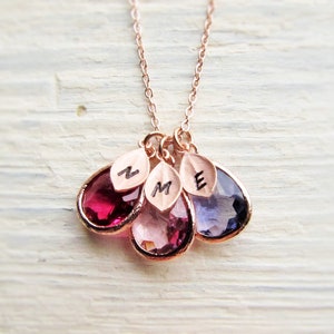 May include: A rose gold necklace with three teardrop-shaped gemstones. The gemstones are red, pink, and purple. Each gemstone has a small gold leaf charm with an initial engraved on it. The initials are N, M, and E.