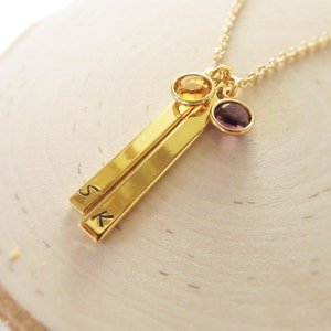 May include: Gold necklace with two gold bars engraved with the letters "S" and "K". The necklace has a purple and a yellow gemstone attached.