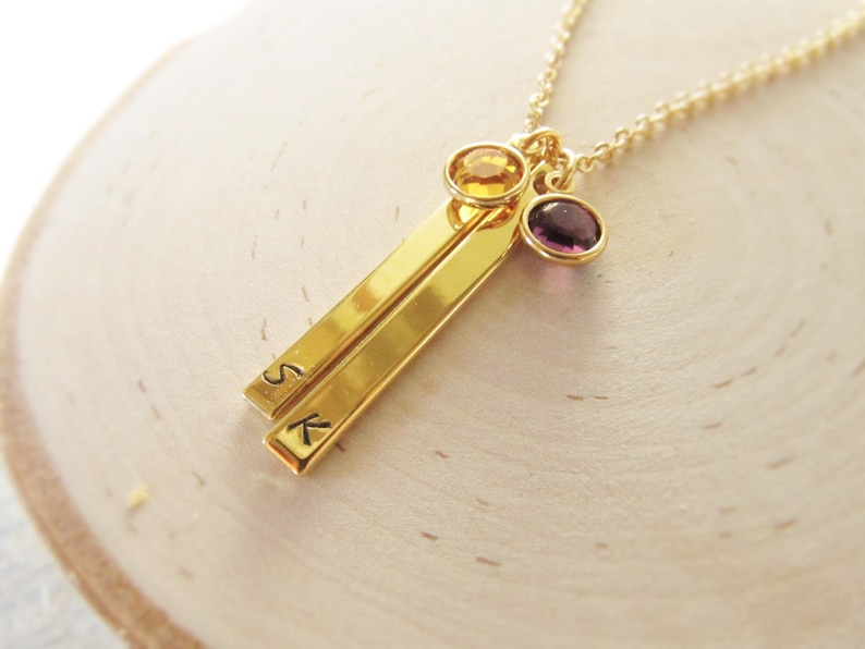 May include: Gold necklace with two gold bars engraved with the letters "S" and "K". The necklace has a purple and a yellow gemstone attached.
